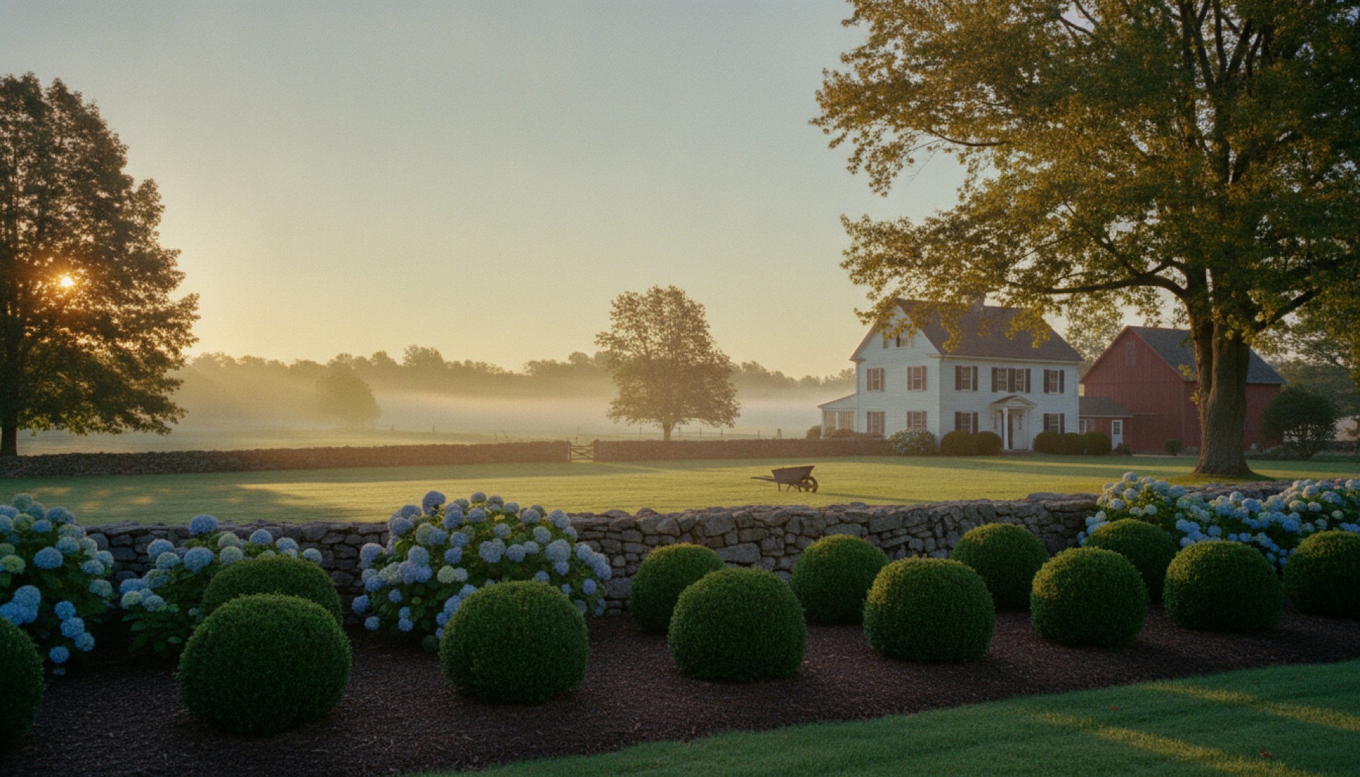 Well-kept country property at early morning with freshly edged mulch beds and clipped boxwood beside an old fieldstone wall and a white farmhouse